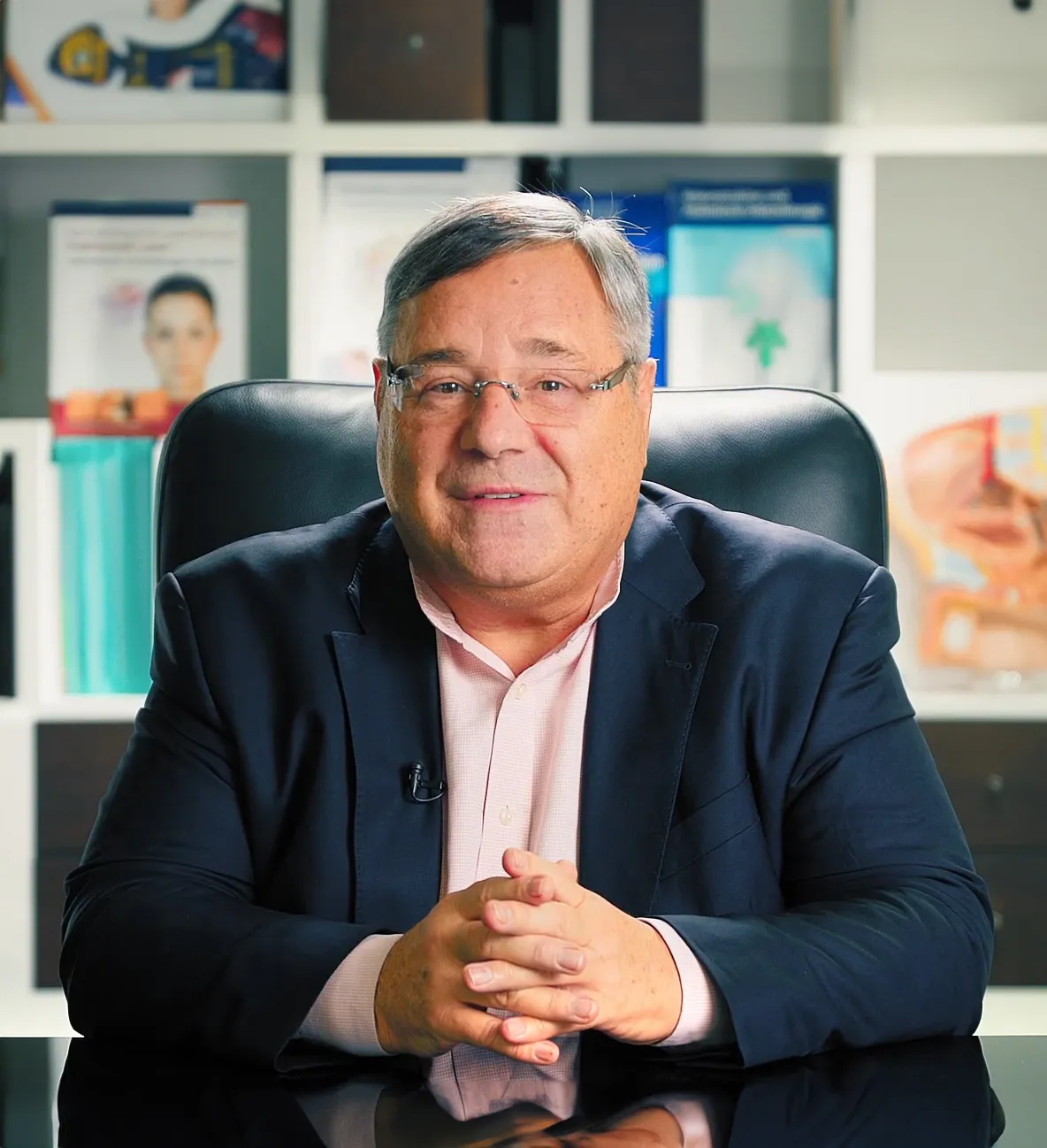 Dr. Deniel Kish, ästhetischer Chirurg in Zürich, lächelt im Büro, umgeben von medizinischen Informationsmaterialien über plastische Chirurgie.