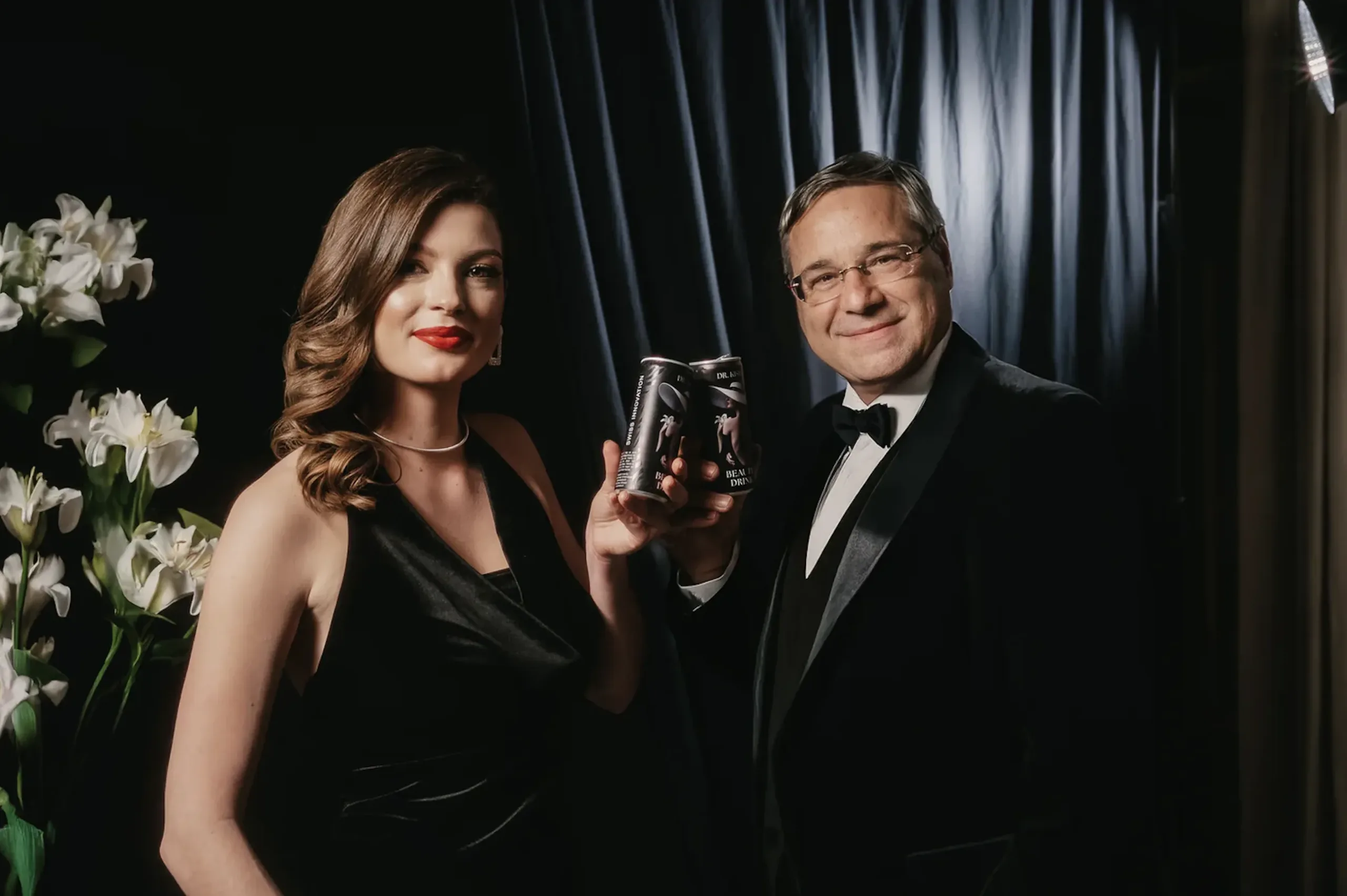 Dr. Kish und eine Frau in eleganter Abendgarderobe halten zusammen Dr. Kish's Age-Reversing Drink in Dosen, umgeben von weißen Lilien und einem dunklen Hintergrund.